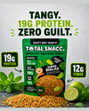 A promotional product shot for Total Snacc Super Bhujia snack packets. The green and black bag is centered on a textured light gray background, with loose brown snack pieces spilled in front, lime halves, mint leaves, and callout badges highlighting 19g protein and 12g fibre.